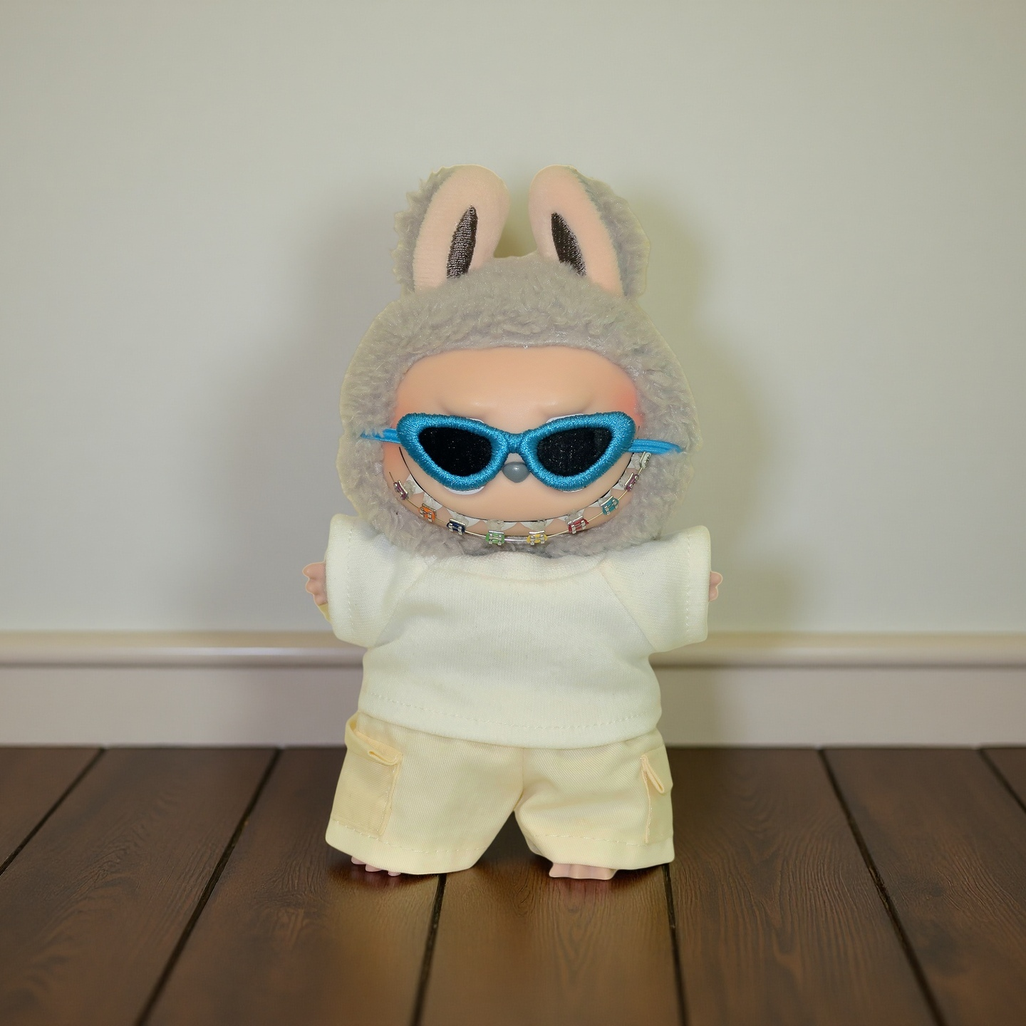 Labubu doll wearing a gray bunny hat, cream casual outfit, blue cyber-style sunglasses, and a colorful beaded eyeglass chain, standing on a wooden floor. Doll not included.