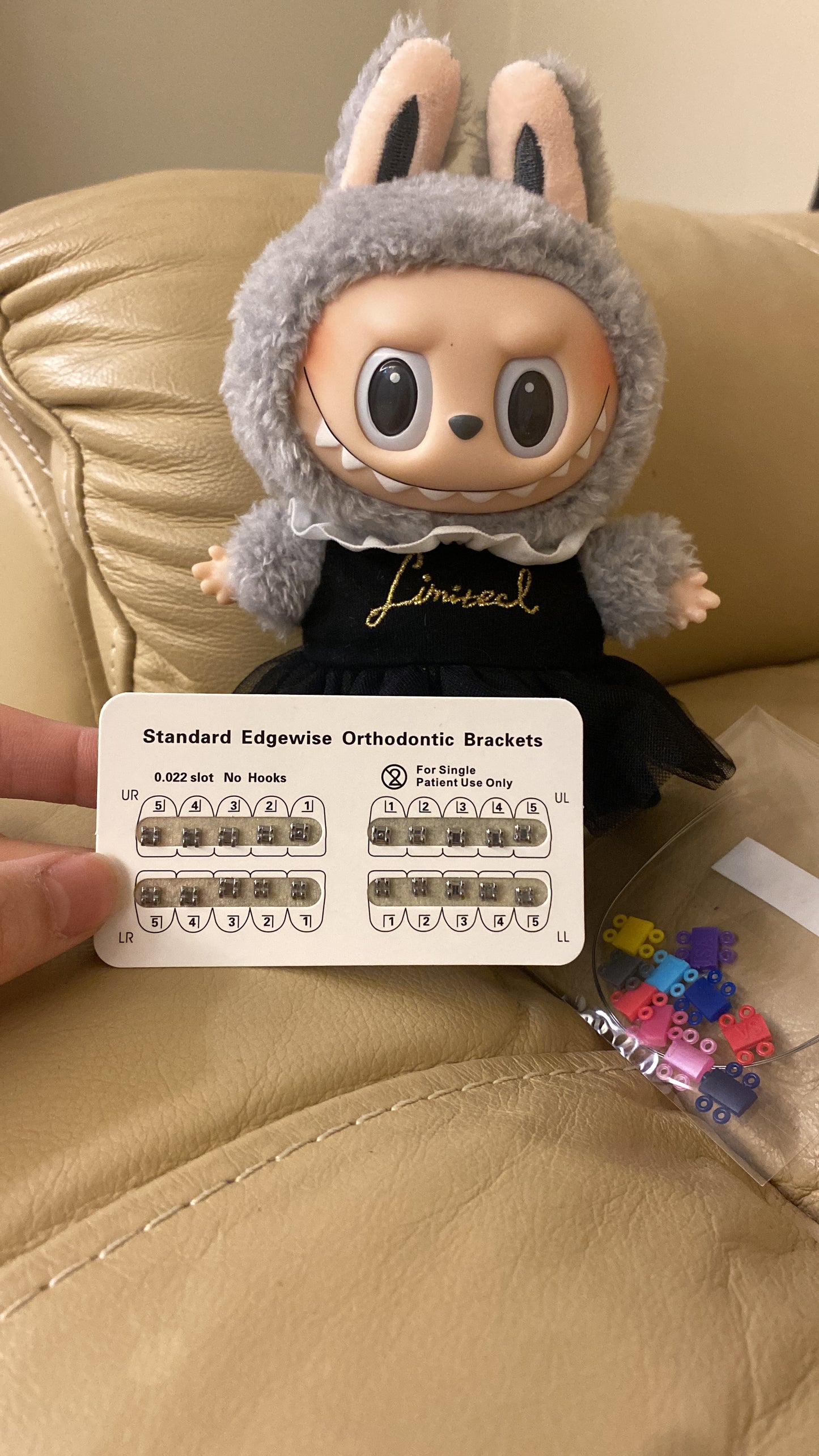 Labubu doll in bunny costume sitting on a beige couch with an orthodontic bracket reference chart and colorful DIY braces parts displayed in the foreground.
