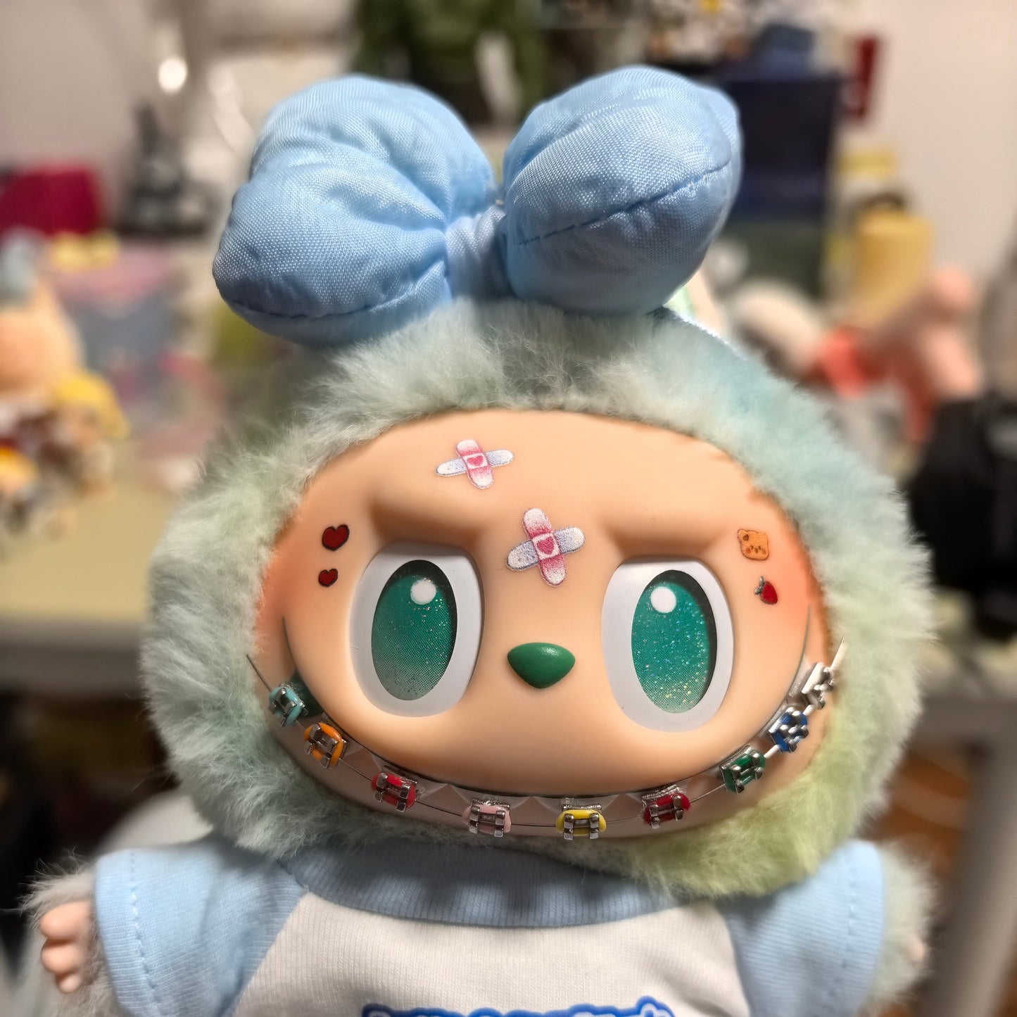 Labubu doll with blue bow and colorful handmade braces, wearing a white T-shirt with Chinese text and dressed in pastel tones. Held in hand with a decorated background. Doll not included.