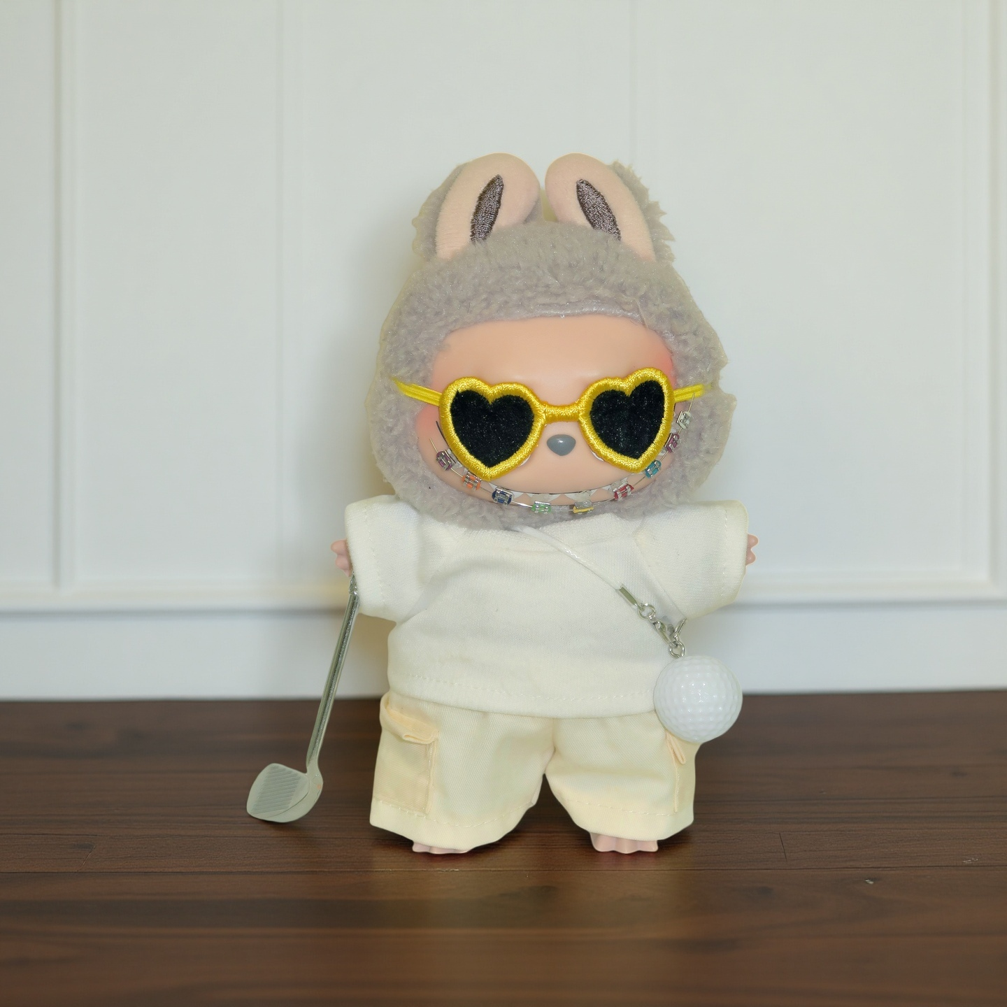 Labubu doll dressed in a cream bunny hat and casual outfit, wearing yellow heart-shaped sunglasses, holding a golf club with a golf ball charm necklace. Doll not included.