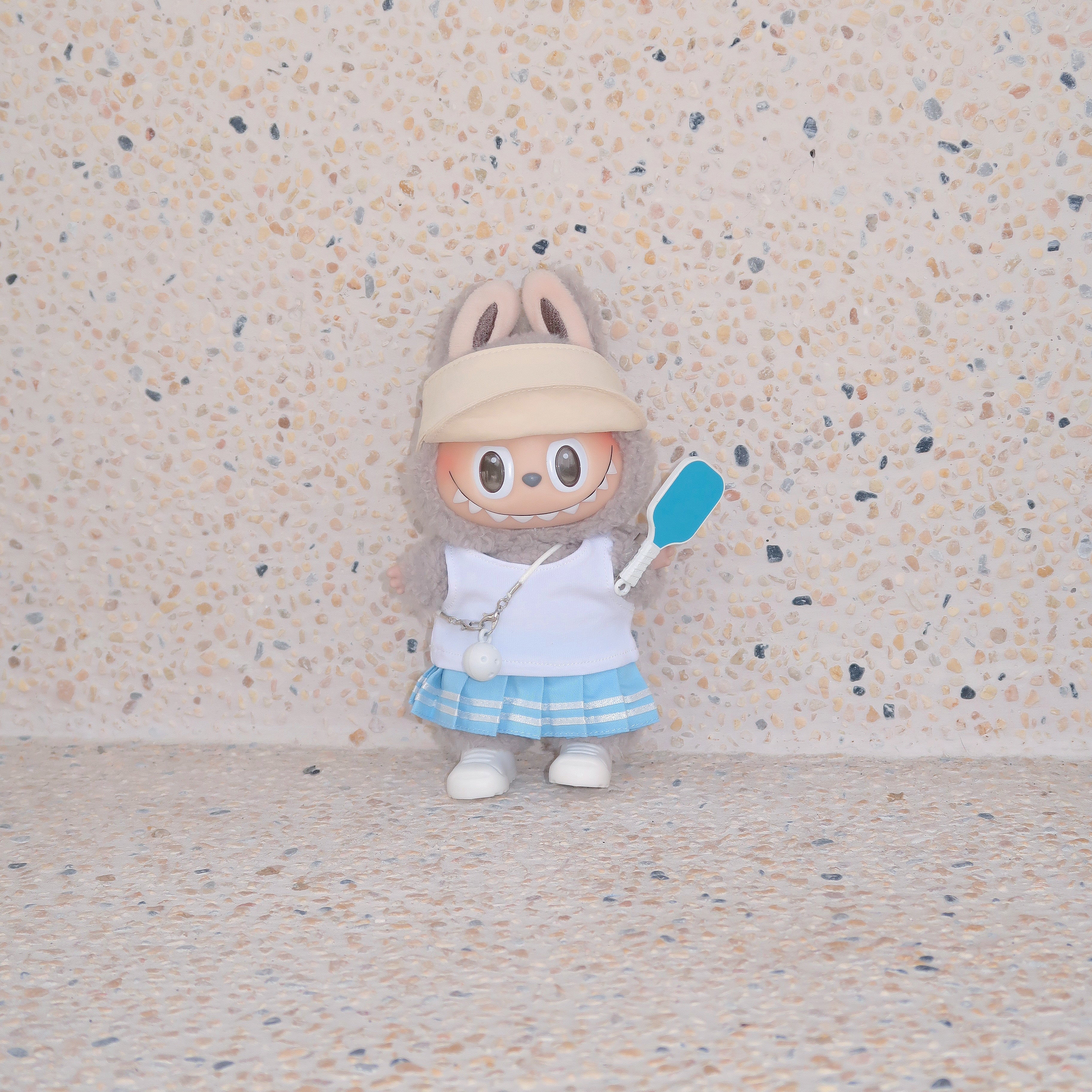 Labubu doll in blue pickleball outfit with khaki visor and paddle, standing against terrazzo beige background, Popanion handmade
