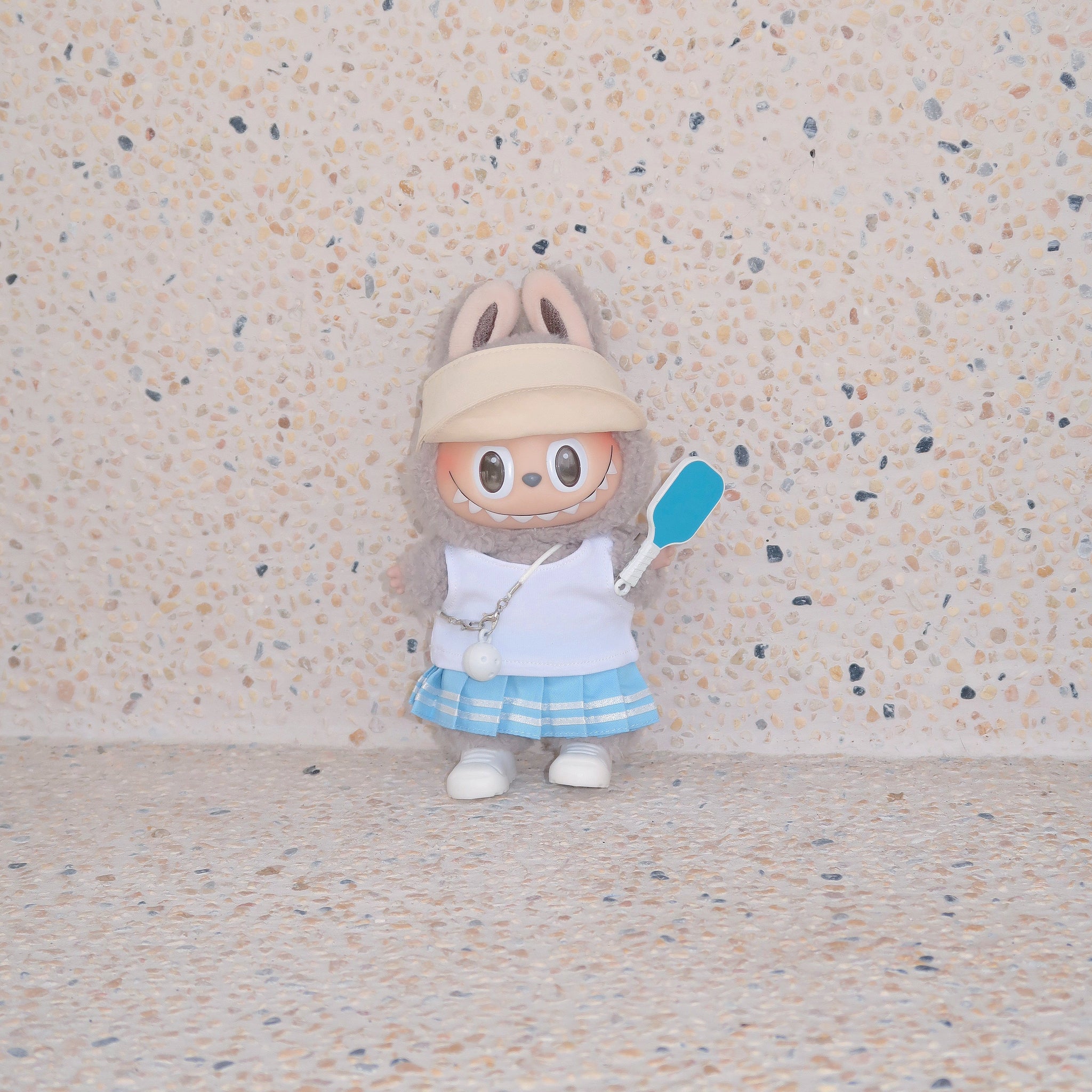 Labubu doll in blue pickleball outfit with khaki visor and paddle, standing against terrazzo beige background, Popanion handmade
