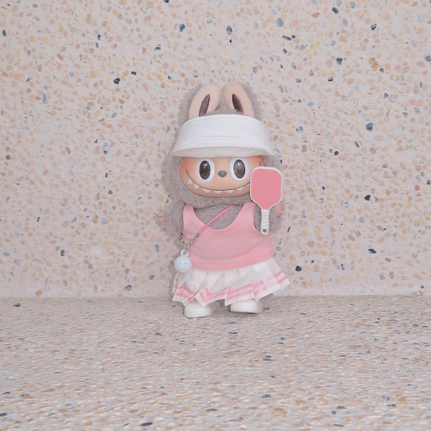 Labubu doll in pink pickleball outfit with visor and paddle, standing against terrazzo beige background, Popanion handmade doll fashion